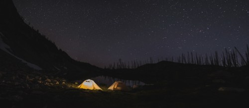 Two tents glow under a starry night sky, reflecting in a calm lake surrounded by dark mountains and trees.