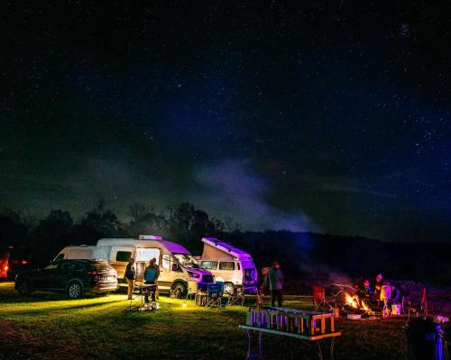 A campsite at night with campers gathered around a fire, surrounded by RVs and a starry sky.