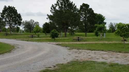 A gravel path winds through a park with trees and picnic tables scattered across a grassy area under a cloudy sky.