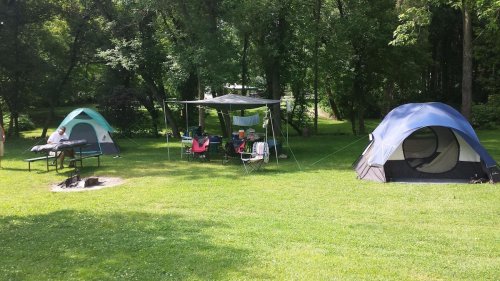 Two tents set up on green grass, with a picnic table and a shaded area nearby, surrounded by trees.