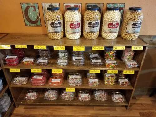 A wooden shelf filled with jars of popcorn and various candy types, labeled with names like "Milk Duds" and "Skittles."