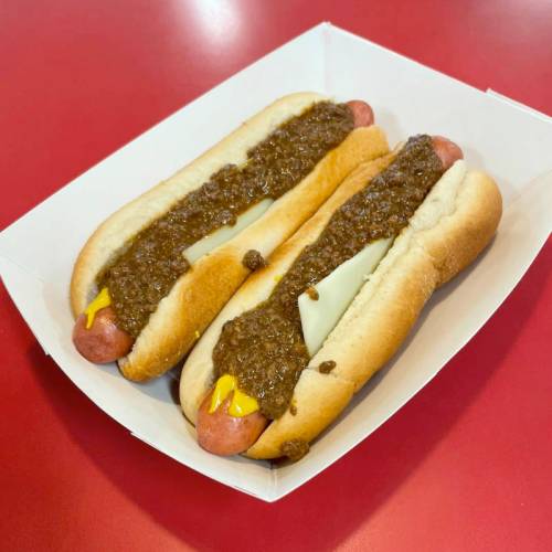 Two hot dogs in a white tray, topped with chili and mustard, on a red table.