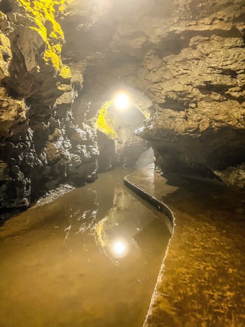 A dimly lit cave with a winding path beside a reflective water surface and rocky walls.