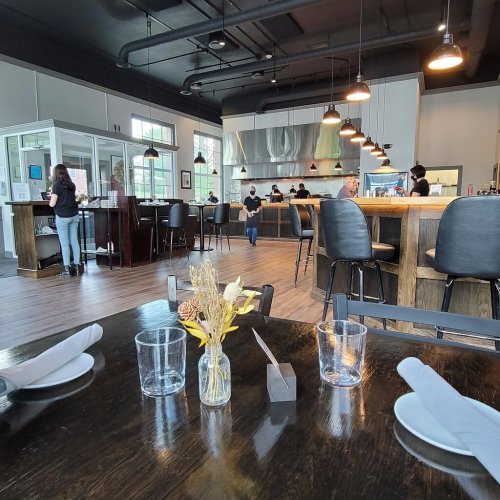 A modern restaurant interior with a bar, kitchen, and tables set for dining, featuring staff and patrons.
