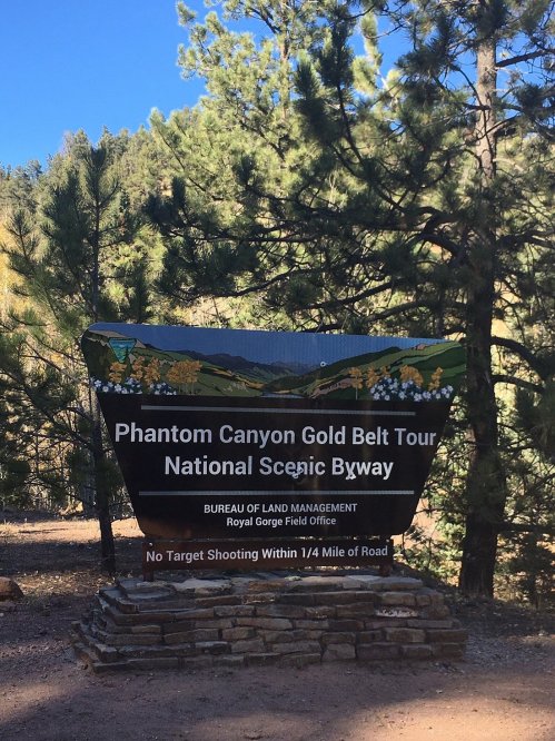 Sign for Phantom Canyon Gold Belt Tour, National Scenic Byway, surrounded by trees and mountains. No target shooting notice.