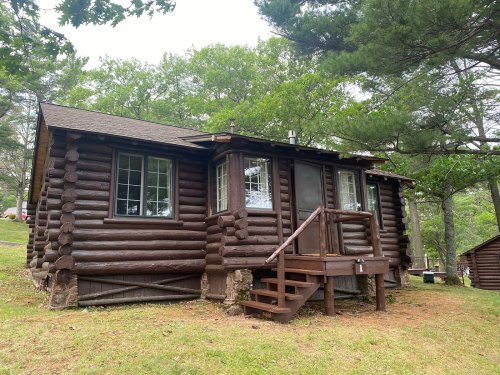 A rustic log cabin with a porch, surrounded by trees and greenery.