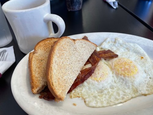A plate with two fried eggs, crispy bacon, and two slices of toast, alongside a white coffee mug.