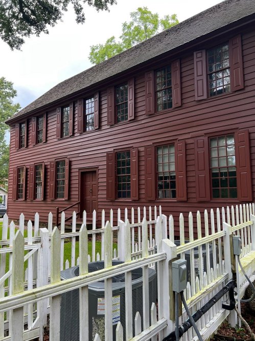A red wooden house with multiple windows, surrounded by a white picket fence and greenery in the background.