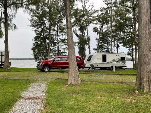 A red truck parked next to a white travel trailer by a lake, surrounded by trees and grassy area.