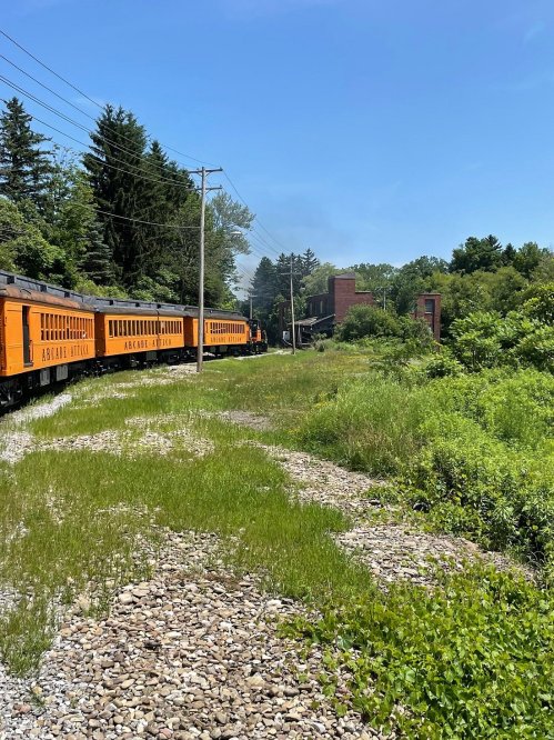 A vintage orange train travels along a grassy track, surrounded by trees and a brick building in the background.