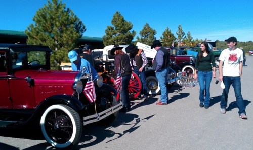 A group of people admire vintage cars decorated with flags at an outdoor event under a clear blue sky.