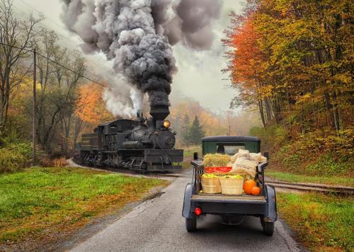 A vintage steam train billows smoke while passing a classic truck loaded with produce on a scenic autumn road.