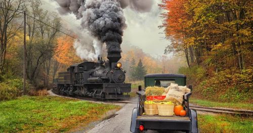 A vintage steam train puffs smoke as it passes a truck loaded with baskets of produce in a colorful autumn landscape.