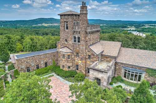 Aerial view of a large stone mansion surrounded by greenery and hills under a partly cloudy sky.