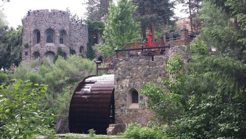 A scenic view of a stone building with a water wheel, surrounded by lush greenery and trees.
