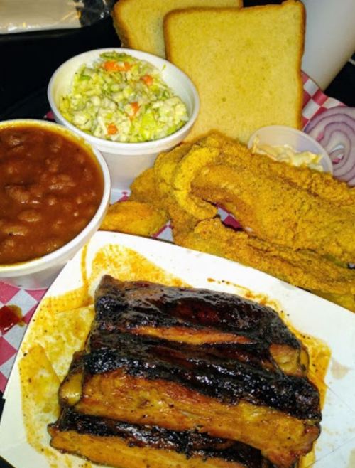 A plate of barbecue ribs, fried catfish, coleslaw, baked beans, and slices of bread on a checkered tablecloth.