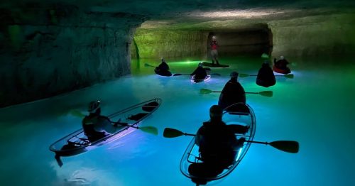 A group of kayakers with glowing paddles exploring a dimly lit cave with turquoise water.