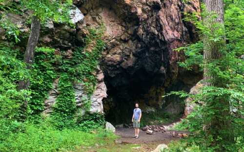 A person stands near a large rock cave surrounded by lush greenery and trees.