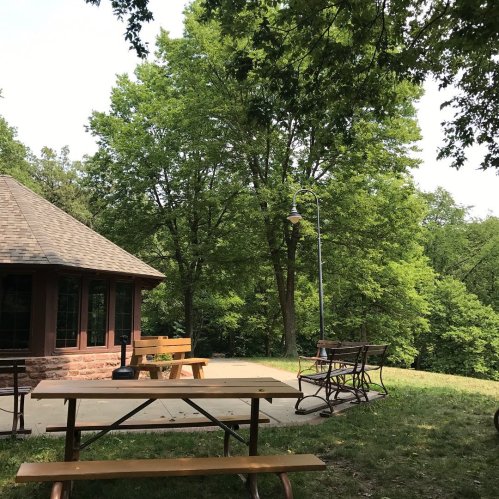 A picnic area with wooden tables near a rustic building, surrounded by lush green trees and a lamp post.