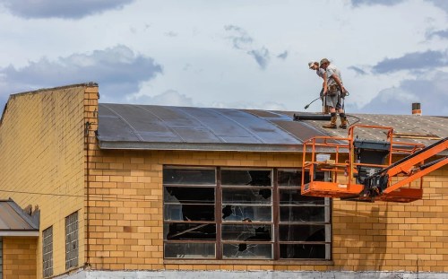 Two workers on a roof using a lift to repair a building with broken windows under a cloudy sky.