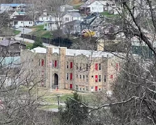 A stone building with red shutters, surrounded by trees and houses on a hillside.