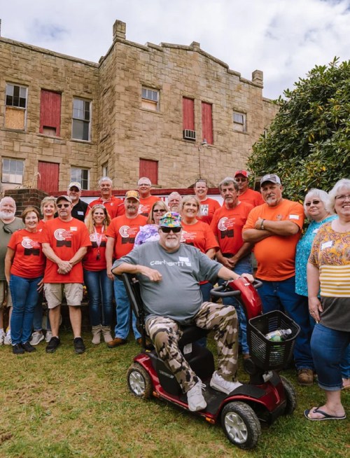 A group of people in red shirts poses together outside a stone building, with one person on a mobility scooter in front.