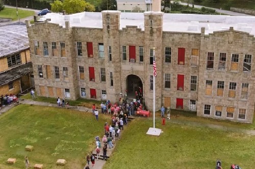 Aerial view of a stone building with red shutters, people gathered outside, and a flagpole in front.