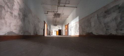 A dimly lit hallway with peeling walls and open doorways, creating an eerie, abandoned atmosphere.