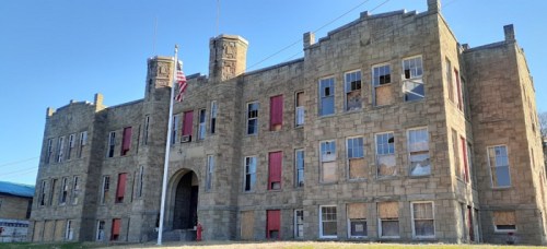 Abandoned stone building with boarded windows and a flagpole in front, set against a clear blue sky.