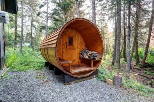 A wooden barrel-shaped structure in a forest, with a small window and stacked firewood inside.