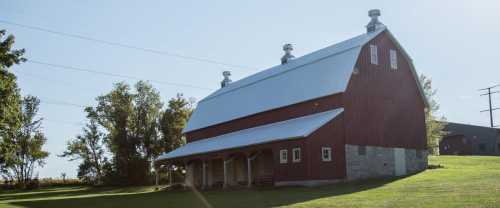 A red barn with a metal roof stands on a grassy field, surrounded by trees and power lines under a clear blue sky.