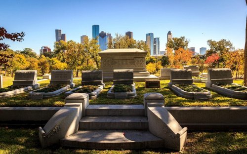 A cemetery with gravestones in the foreground and a city skyline in the background under a clear blue sky.