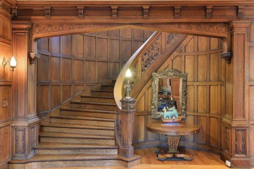 Elegant wooden staircase with intricate carvings, a decorative mirror, and a round table in a classic interior.