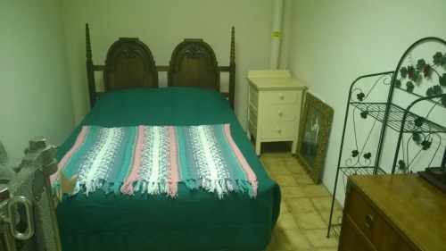 A simple bedroom featuring a double bed with a green cover, a nightstand, and a framed picture on the wall.