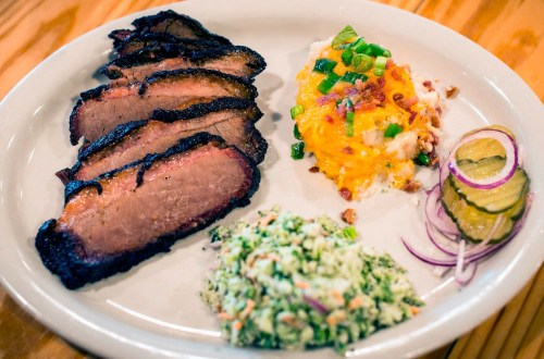 A plate featuring sliced smoked brisket, coleslaw, cheesy potatoes, pickles, and red onion.