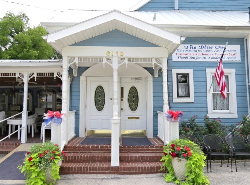 A blue building with a white porch, celebrating its 30th anniversary, adorned with flowers and an American flag.