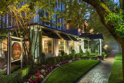 A charming inn with a welcoming entrance, surrounded by lush gardens and colorful flowers, illuminated at dusk.