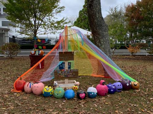 A colorful display of painted pumpkins arranged under a rainbow canopy in a park setting.