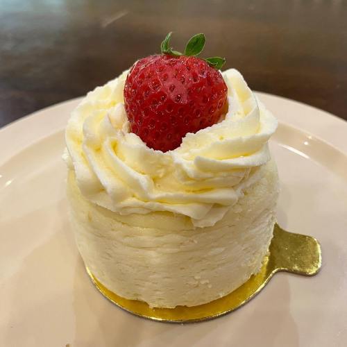 A small cheesecake topped with whipped cream and a fresh strawberry, sitting on a gold base on a white plate.