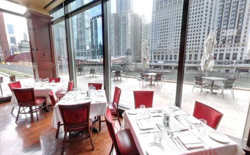 A restaurant with red chairs and white tablecloths, featuring large windows overlooking a cityscape and river.