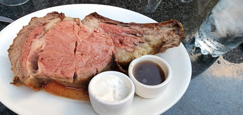 A large, juicy prime rib served on a plate with two dipping sauces and a glass of water in the background.