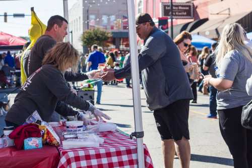 A busy street fair scene with vendors serving food to customers under a tent, surrounded by people enjoying the event.