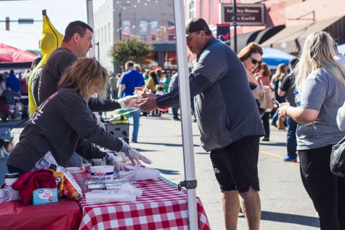 A busy street fair scene with vendors serving food to customers under a tent, surrounded by people enjoying the event.