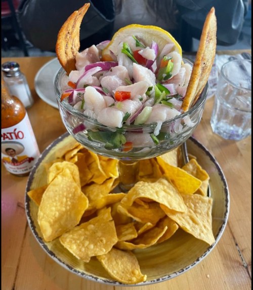 A glass bowl of ceviche with fish, onions, and herbs, served with tortilla chips and a slice of lemon.