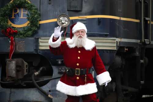 Santa Claus waves beside a decorated train, wearing his classic red suit and hat, with a festive wreath in the background.