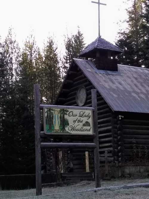 Log cabin church with a cross on the roof and a sign reading "Our Lady of the Woodland," surrounded by trees.