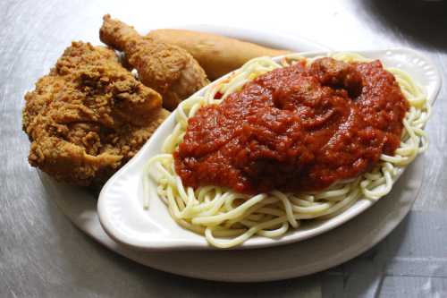 A plate of spaghetti with marinara sauce, fried chicken, and a piece of cornbread on a silver surface.