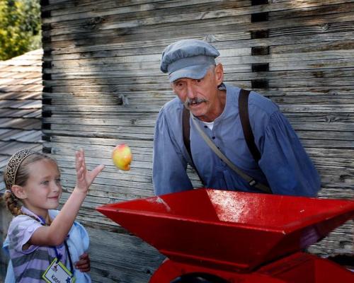 A young girl tosses an apple to an older man beside a red apple press, set against a rustic wooden backdrop.