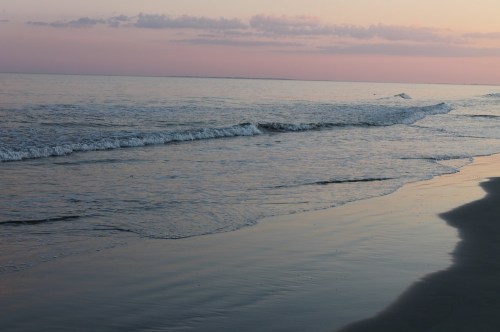 Gentle waves lapping at a serene beach during sunset, with soft pink and purple hues in the sky.
