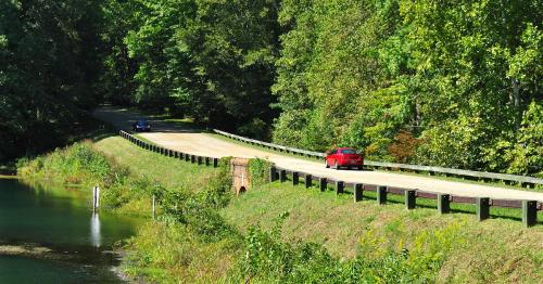A red car drives along a winding road beside a lush green forest and a calm body of water.